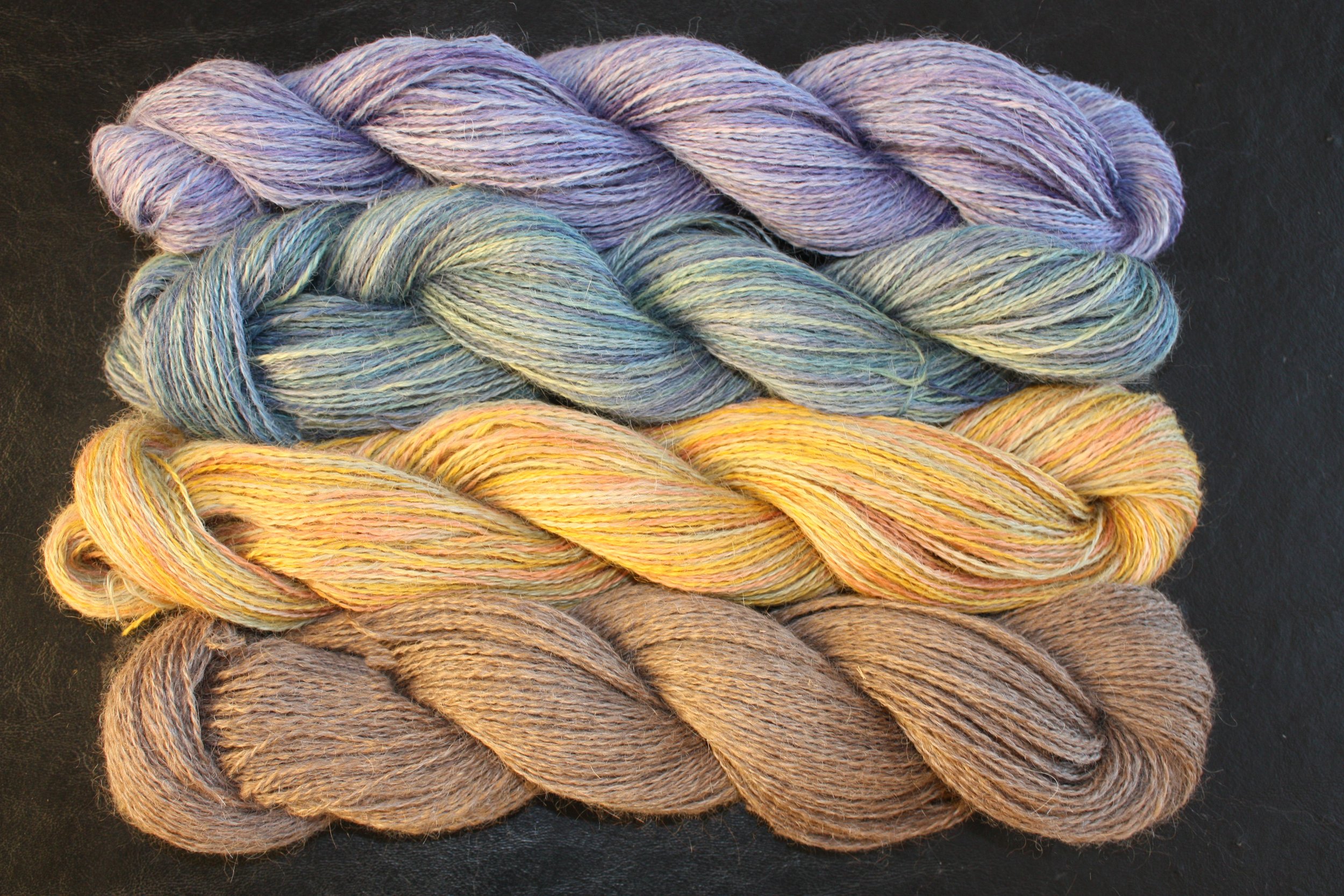 Jill+Knuckles+-+1_Product_Hand+Dyed+Yarn+1 Four skeins of hand-dyed yarn in soft pastel colors, including lavender, teal, yellow, and brown, arranged horizontally against a dark background, showcasing the texture and quality of the fibers for knitting and crafting projects.